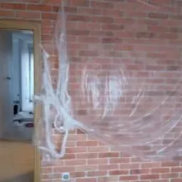 Ściana z cegły pokryta folią ochronną podczas prac remontowych, widoczne gniazdko elektryczne i fragment otwartego przejścia do innego pomieszczenia.