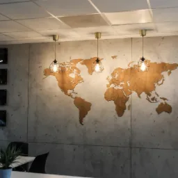 Biuro z mapą świata wyciętą z drewna na szarej ścianie, oświetloną trzema lampami wiszącymi w metalowych kloszach. Na ścianie po lewej stronie wiszą cztery czarno-białe zdjęcia w ramkach.