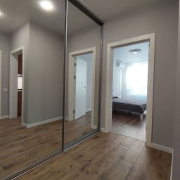 RP Work Sp. z o.o. - Nowoczesne wnętrze mieszkania z drewnopodobną podłogą, szarą kolorystyką ścian i zabudowaną szafą z przesuwnymi drzwiami lustrzanymi, ukazujące pokój z łóżkiem w tle.