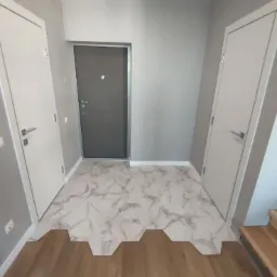 Wykończenie przedpokoju z heksagonalnymi płytkami imitującymi marmur, przechodzącymi w drewnianą podłogę, widoczne dwie białe drzwi i szare drzwi wejściowe.