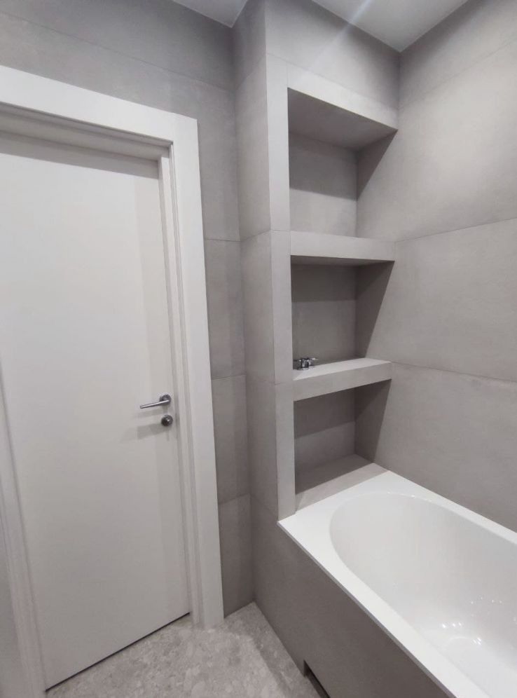 Wykończona łazienka w odcieniach szarości z wanną i wnękami w ścianie, widoczna biała stolarka drzwiowa.