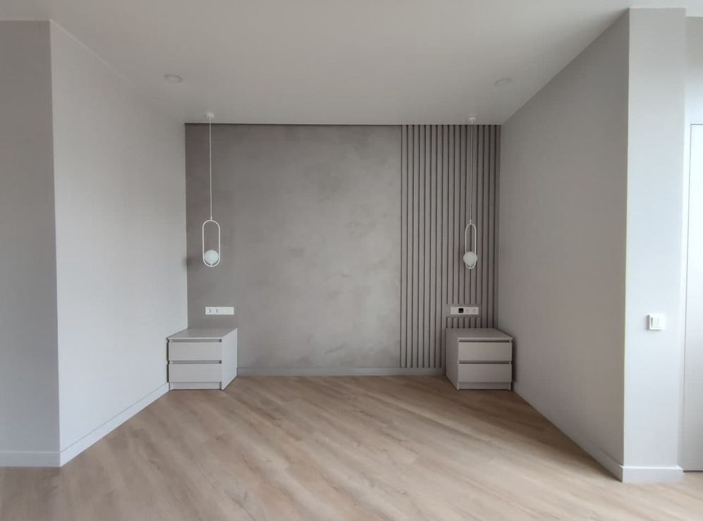Minimalistyczna sypialnia z szarą ścianą, drewnianą podłogą, białymi szafkami nocnymi i wiszącymi lampami o owalnym kształcie.
