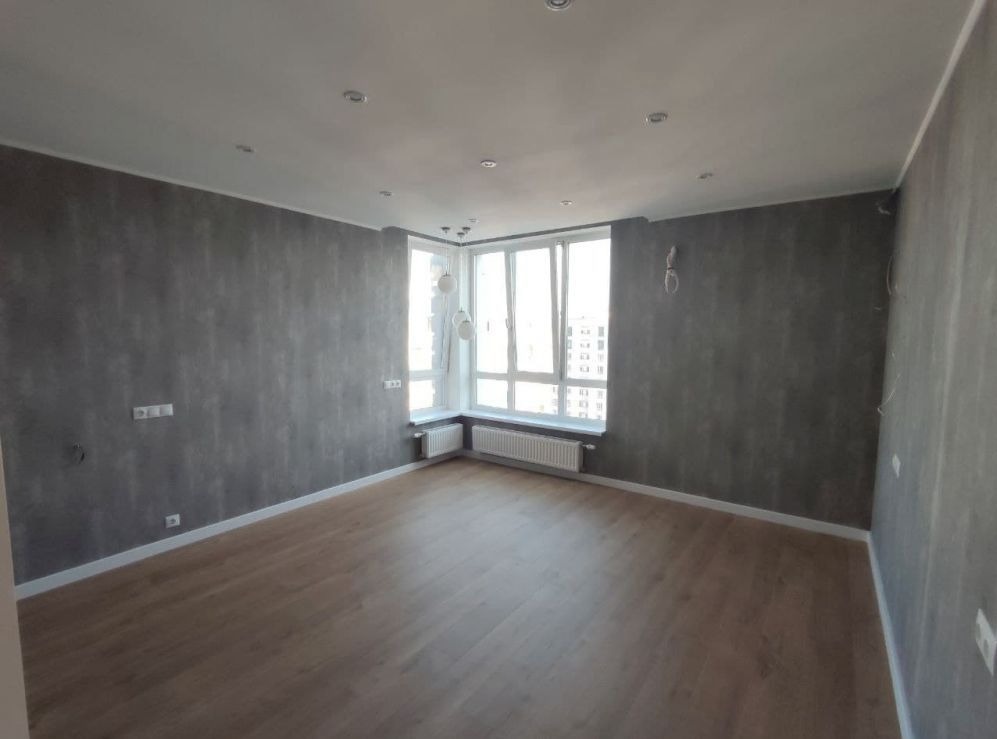 Pusty pokój po remoncie z szarymi ścianami o fakturze betonu, parkietem i dużym oknem narożnym z białymi grzejnikami.