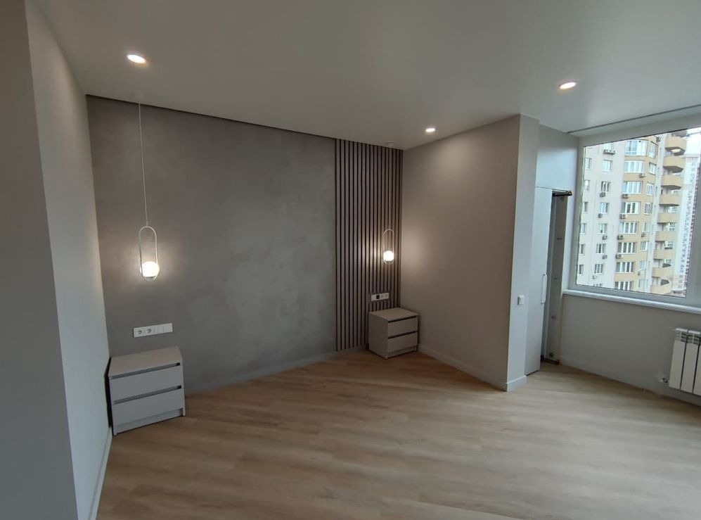 Nowoczesna sypialnia w Ząbkach po remoncie: jasna podłoga, szara ściana z dekoracyjnymi panelami pionowymi, minimalistyczne szafki nocne z lampkami wiszącymi i widok na miasto z okna.