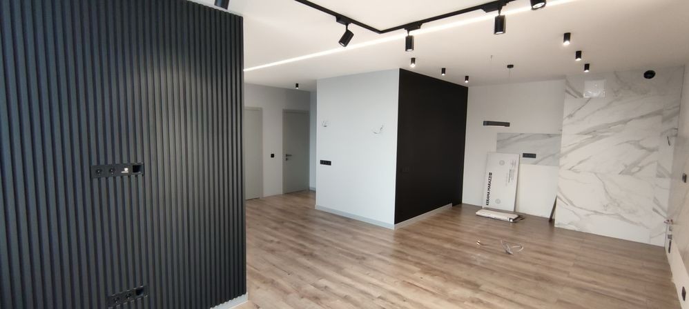 Nowoczesne wnętrze z drewnianą podłogą, czarną ścianą z pionowych paneli z gniazdkami, białymi ścianami z czarnymi akcentami, oświetleniem punktowym na suficie i marmurowymi płytami ułożonymi...