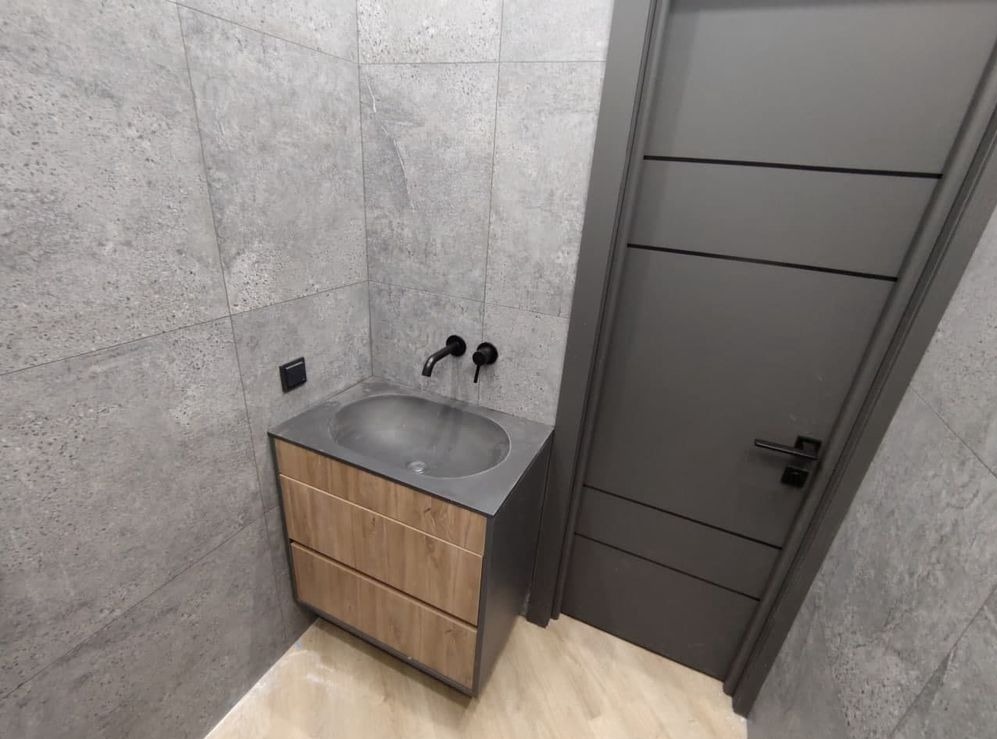 Nowoczesna łazienka z szarą, teksturowaną płytką ścienną, drewnianą szafką pod umywalką z szarą, metalową umywalką i czarną armaturą, oraz szare drzwi z poziomymi frezowaniami.