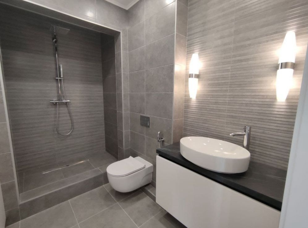 Nowoczesna łazienka w odcieniach szarości z kabiną prysznicową typu walk-in, podwieszaną toaletą i owalną umywalką na białej szafce z czarnym blatem, oświetlona dwoma pionowymi lampami ściennymi.