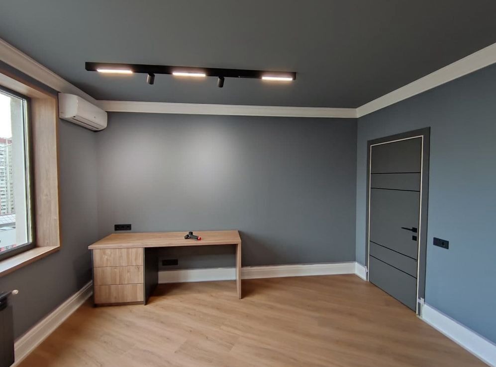 Wnętrze pokoju po remoncie: szare ściany, drewniana podłoga, biurko z szufladami, nowoczesne oświetlenie sufitowe i klimatyzator, widok na okno.