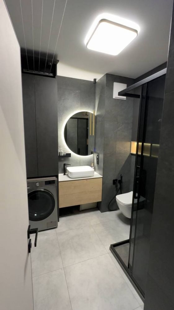 Nowoczesna łazienka z szarymi płytkami, okrągłym lustrem z podświetleniem LED, pralką i kabiną prysznicową z czarnymi ramami.