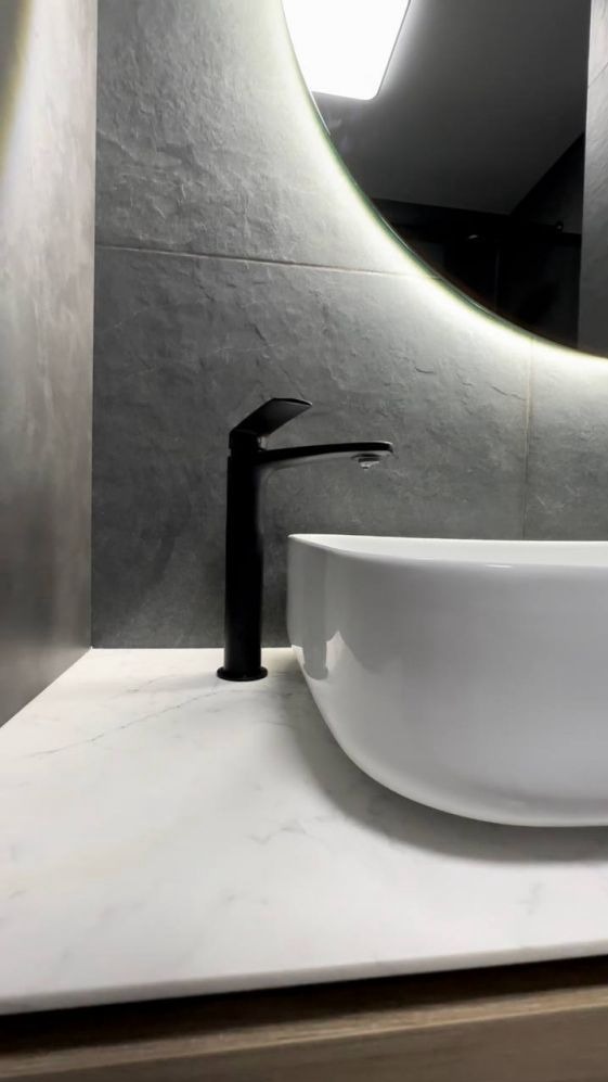 Nowoczesna łazienka z białą umywalką nablatową, czarną armaturą i podświetlanym lustrem w kształcie koła na tle szarej ściany z imitacją kamienia.