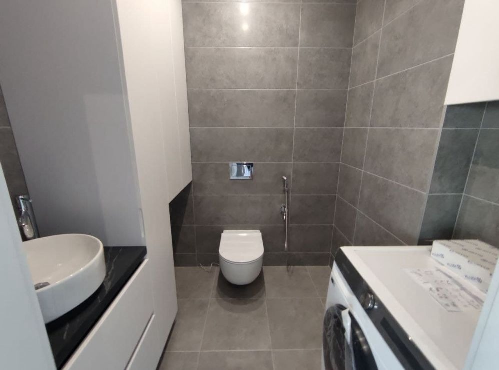 Nowoczesna łazienka z szarymi płytkami, wiszącą miską WC z bidetką, umywalką nablatową na czarnym blacie i pralką.