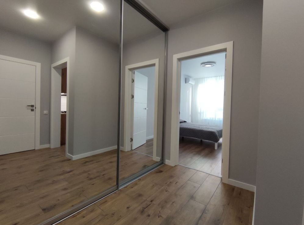 Nowoczesne wnętrze mieszkania z drewnianą podłogą, szarą kolorystyką ścian i zabudowaną szafą z lustrzanymi drzwiami przesuwnymi, widok na sypialnię z łóżkiem.