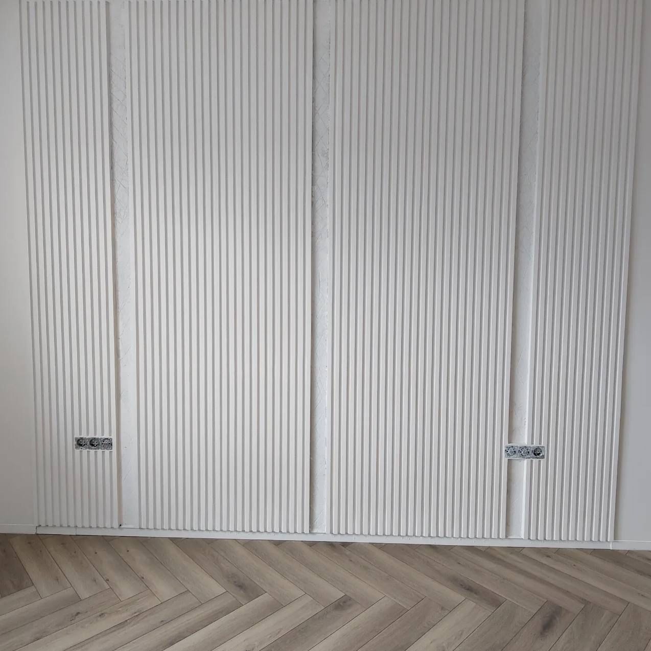 Nowoczesna ściana z białymi panelami dekoracyjnymi z pionowymi żłobieniami, widoczne gniazdka elektryczne i parkiet ułożony w jodełkę.