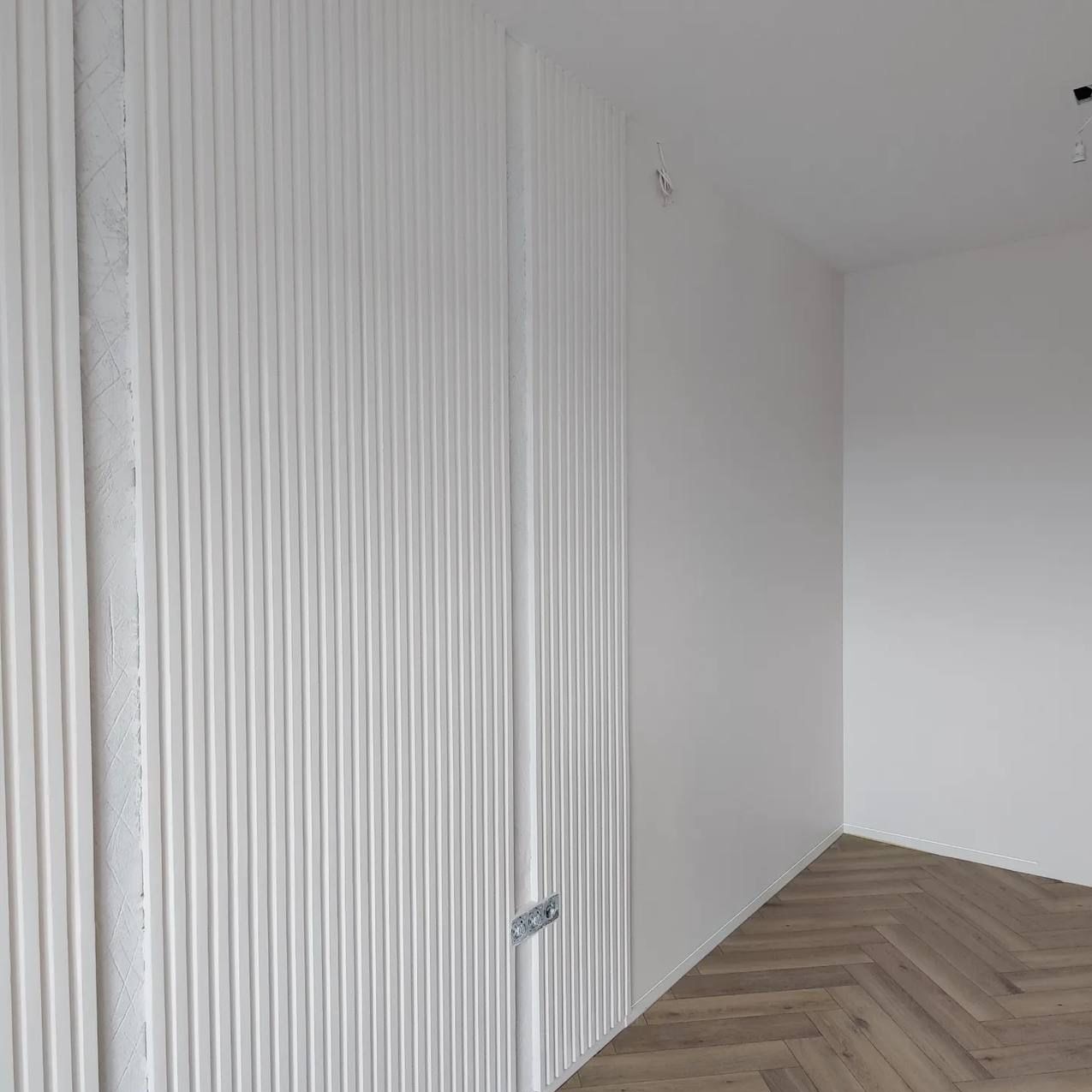 Białe, pionowe lamele dekoracyjne na ścianie z widoczną puszką elektryczną, jasna ściana i podłoga ułożona w jodełkę.