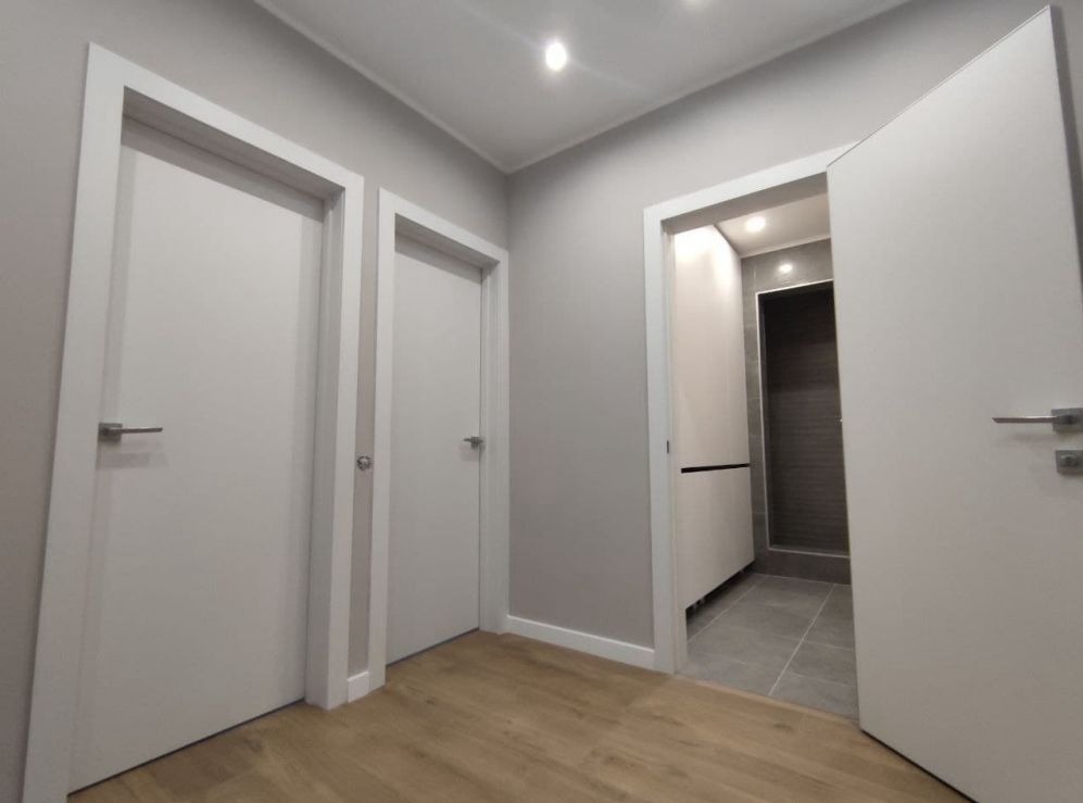 Nowoczesny korytarz z trzema białymi drzwiami z srebrnymi klamkami, podłoga wyłożona panelami, widok na wykończoną łazienkę z szarymi płytkami.