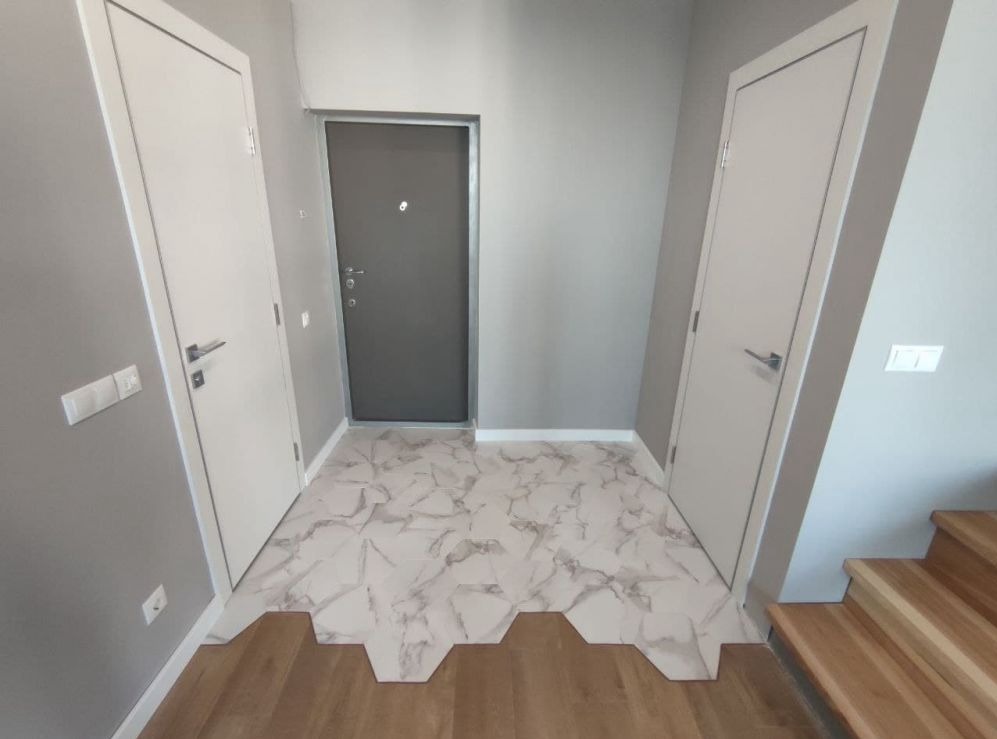 Wykończenie przedpokoju z heksagonalnymi płytkami imitującymi marmur, przechodzącymi w drewnianą podłogę, widoczne dwie białe drzwi i szare drzwi wejściowe.