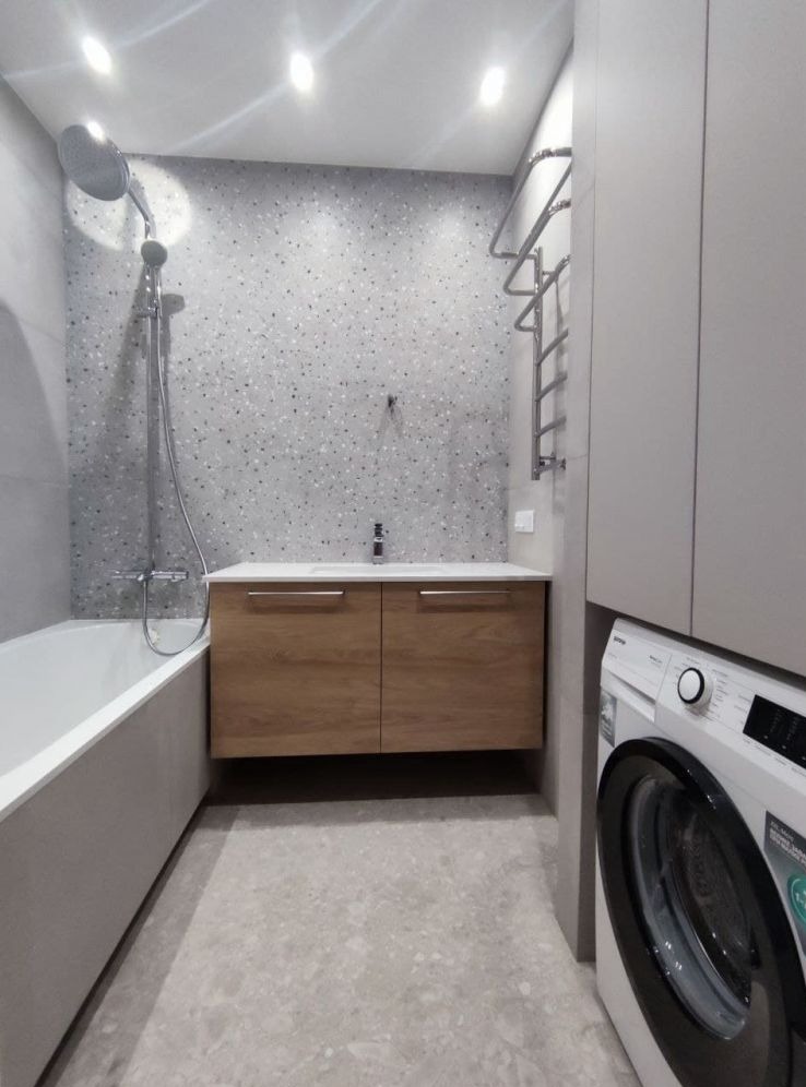 Nowoczesna łazienka z drewnianą szafką pod umywalką, baterią, wanną, prysznicem, grzejnikiem drabinkowym i pralką Gorenje, wykończona płytkami imitującymi lastryko.
