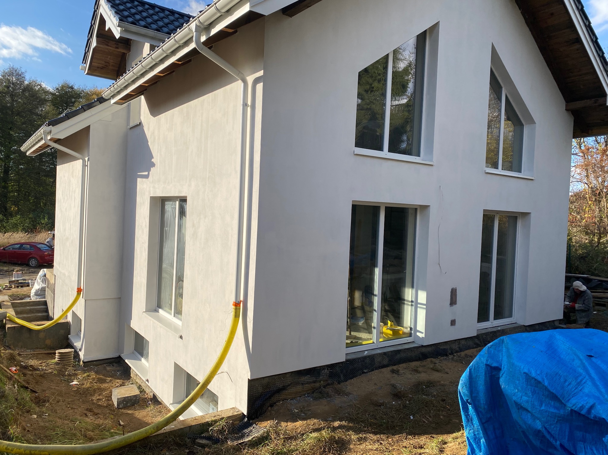 Biały dom z nowoczesnymi oknami, widok z zewnątrz. Żółty wąż leży na ziemi. Prace wykończeniowe w toku, widoczny fragment niebieskiej plandeki i pracownik w tle.