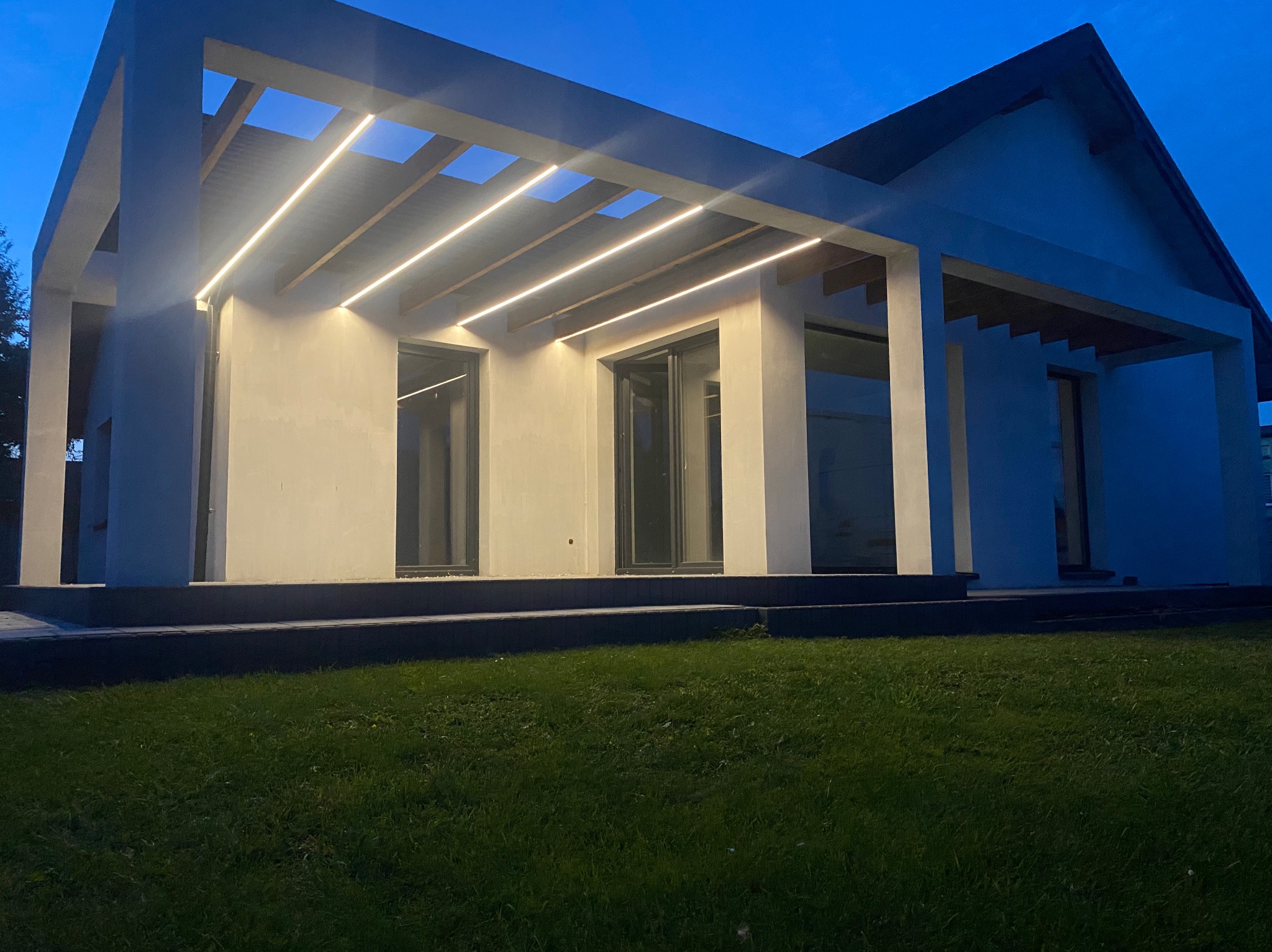 Nowoczesny dom z płaskim dachem i oświetleniem LED wzdłuż krawędzi dachu, widok z trawnika o zmierzchu. Minimalistyczna architektura, ciemne okna.