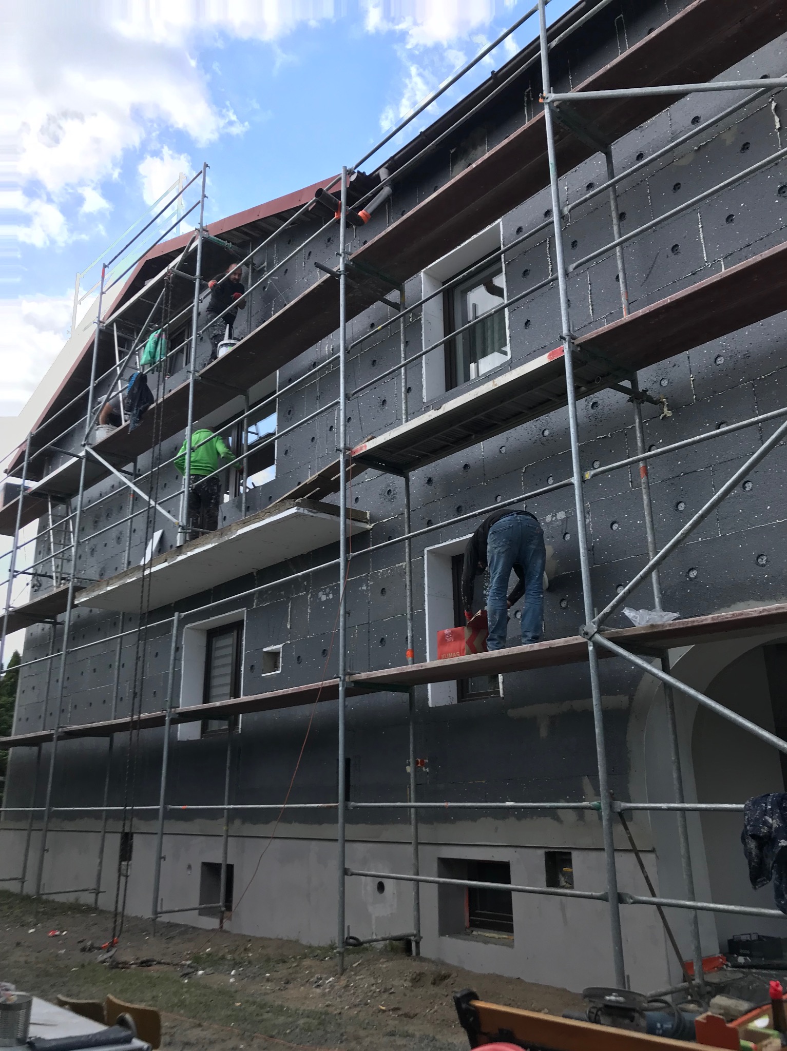 Ocieplanie elewacji domu jednorodzinnego z użyciem rusztowań. Widoczne płyty izolacyjne z charakterystycznymi otworami. Prace prowadzone przez kilku pracowników.