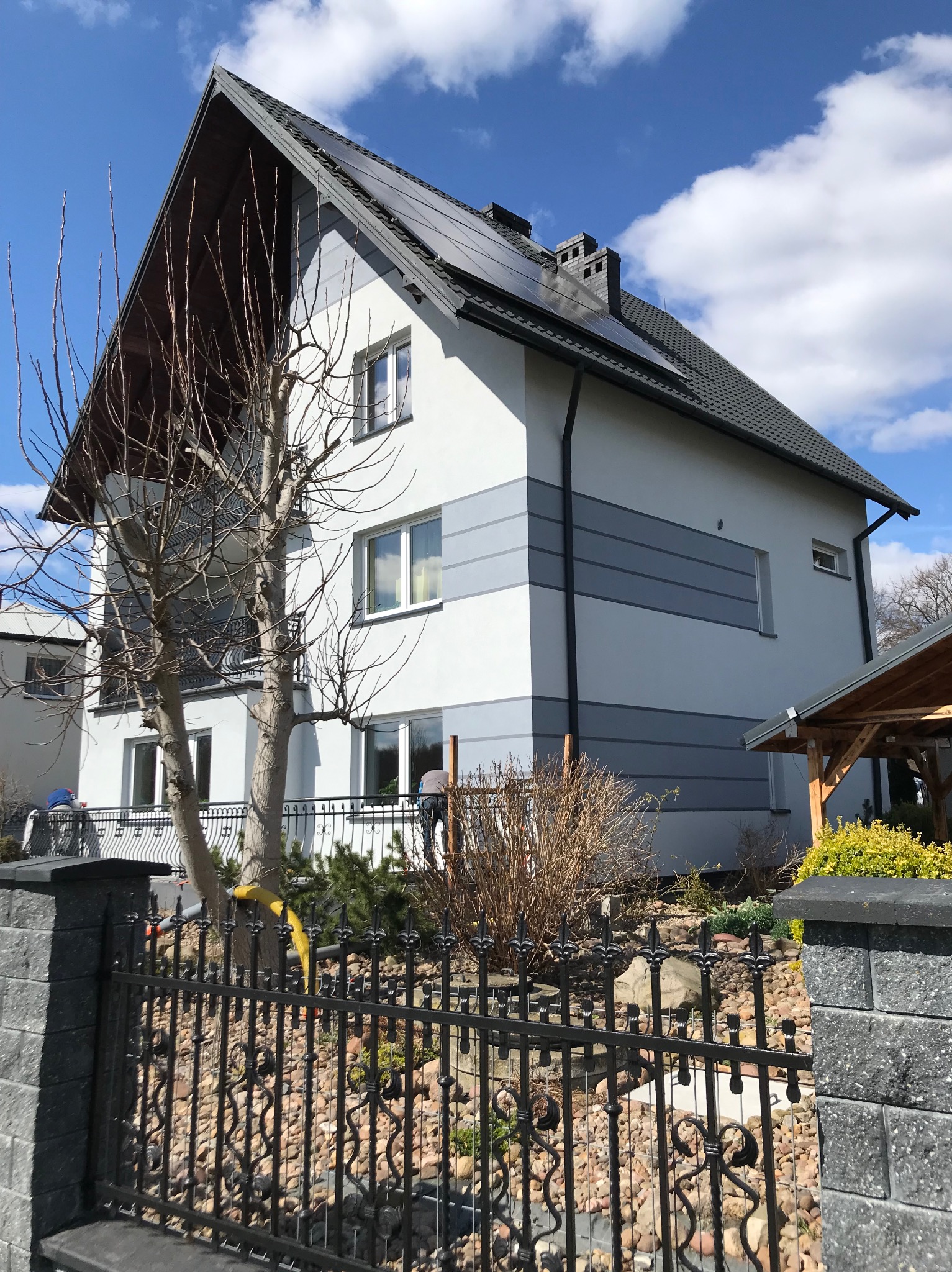 Dwukondygnacyjny dom z szarą elewacją i panelami słonecznymi na dachu, widziany zza ozdobnego, metalowego ogrodzenia na tle błękitnego nieba.
