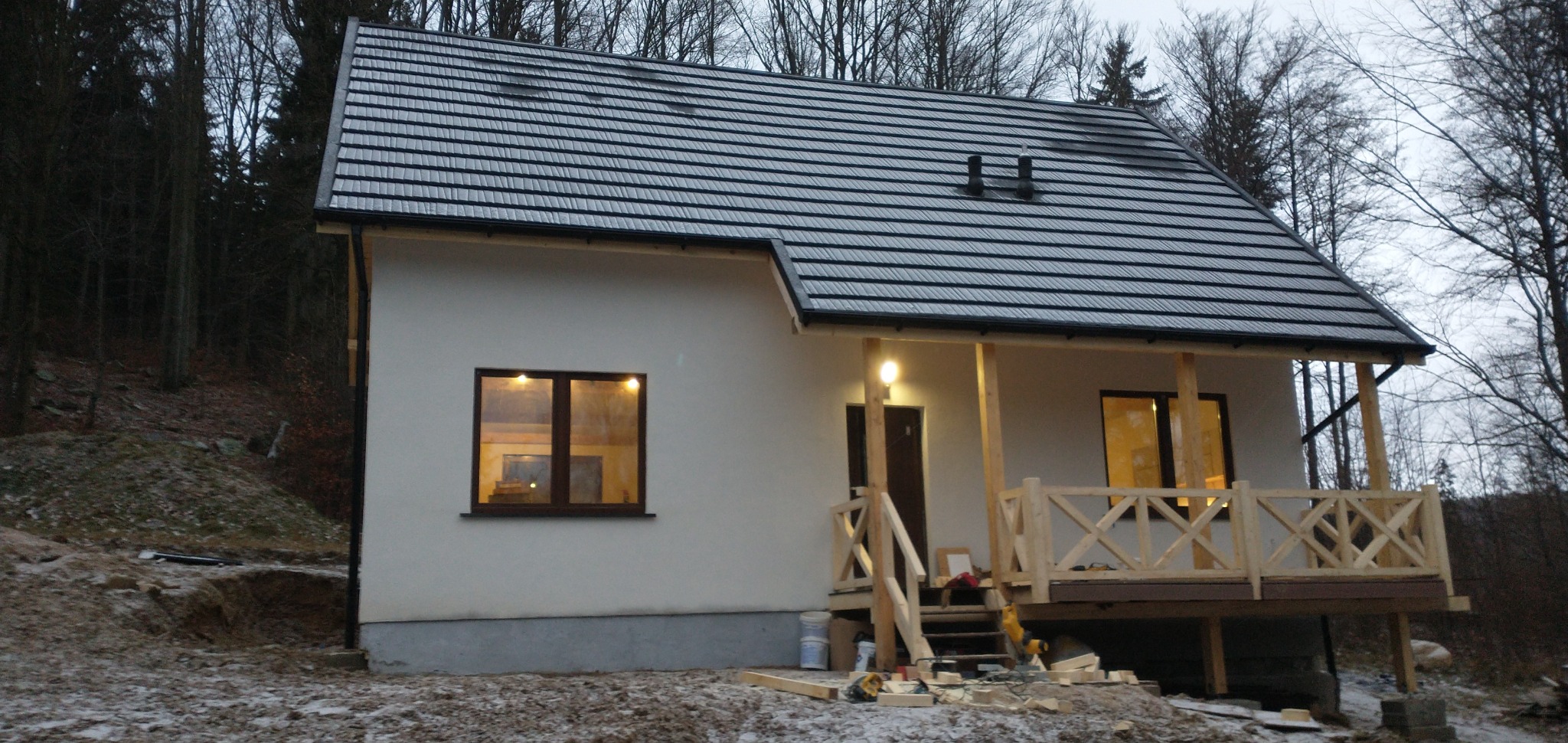 Niedokończony dom z drewnianym gankiem i czarnym dachem na tle lasu. Widoczne narzędzia i materiały budowlane. Okna oświetlone od wewnątrz.