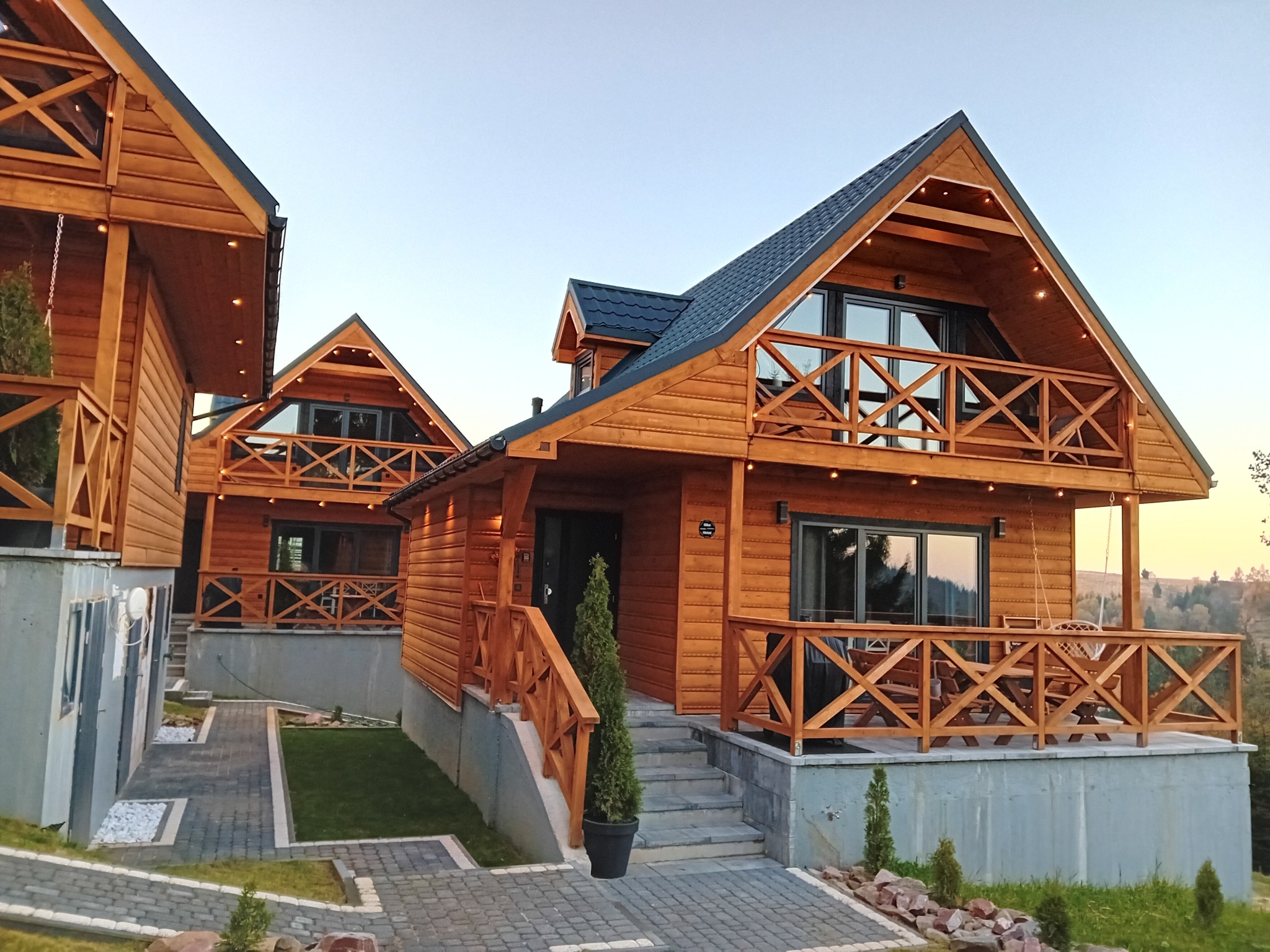 Kompleks domków letniskowych z drewna, z balkonami i schodami, otoczonych zielenią i ścieżkami z kostki brukowej, w tle zalesione wzgórza o zmierzchu.