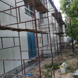 Budynek w trakcie ocieplania elewacji, widoczne rusztowania, zabezpieczone okno folią, oraz przygotowane materiały budowlane na ziemi.