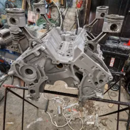 Szary blok silnika V8 z zamontowanymi tłokami na stojaku warsztatowym, widoczne ślady świeżego malowania i otoczenie warsztatowe z narzędziami i przewodami.