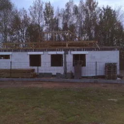 Surowy stan domu jednorodzinnego z białych bloczk&oacute;w, widoczne drewniane elementy konstrukcji dachu, palety z materiałami budowlanymi i ogrodzenie siatkowe.