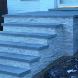 Wykończone schody wejściowe do domu, z szarymi stopniami i obłożone jasną mozaiką kamienną, widoczne niedociągnięcia w dolnej części.