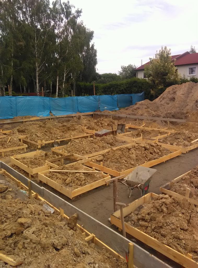 Fundamenty domu w trakcie budowy: drewniane szalunki wypełnione ziemią, częściowo zalane betonem, taczka i łopata, w tle drzewa i dom z czerwonym dachem, zabezpieczenie niebieską plandeką.
