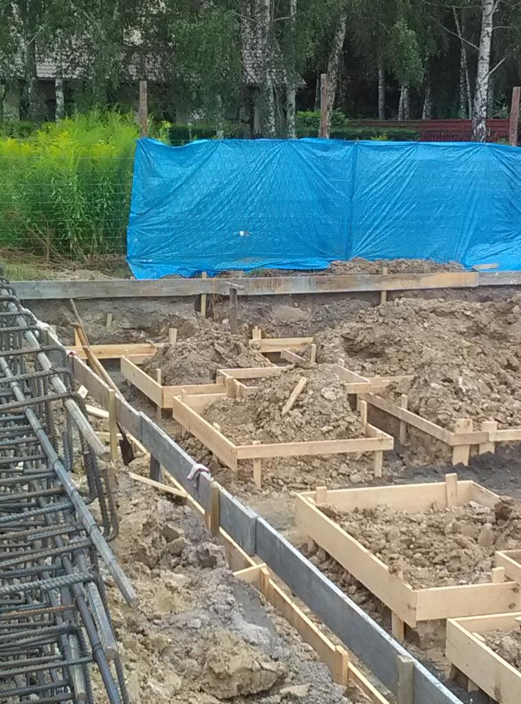Przygotowanie terenu pod fundamenty domu z drewnianymi szalunkami i zbrojeniem stalowym, w tle niebieska plandeka i drzewa.
