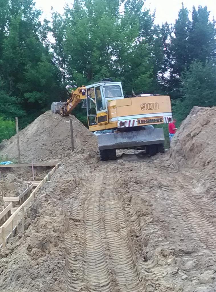 Żółta koparka Liebherr 900 na placu budowy, z widocznymi śladami opon na ziemi i drewnianym szalunkiem fundamentów w tle, drzewa w tle.