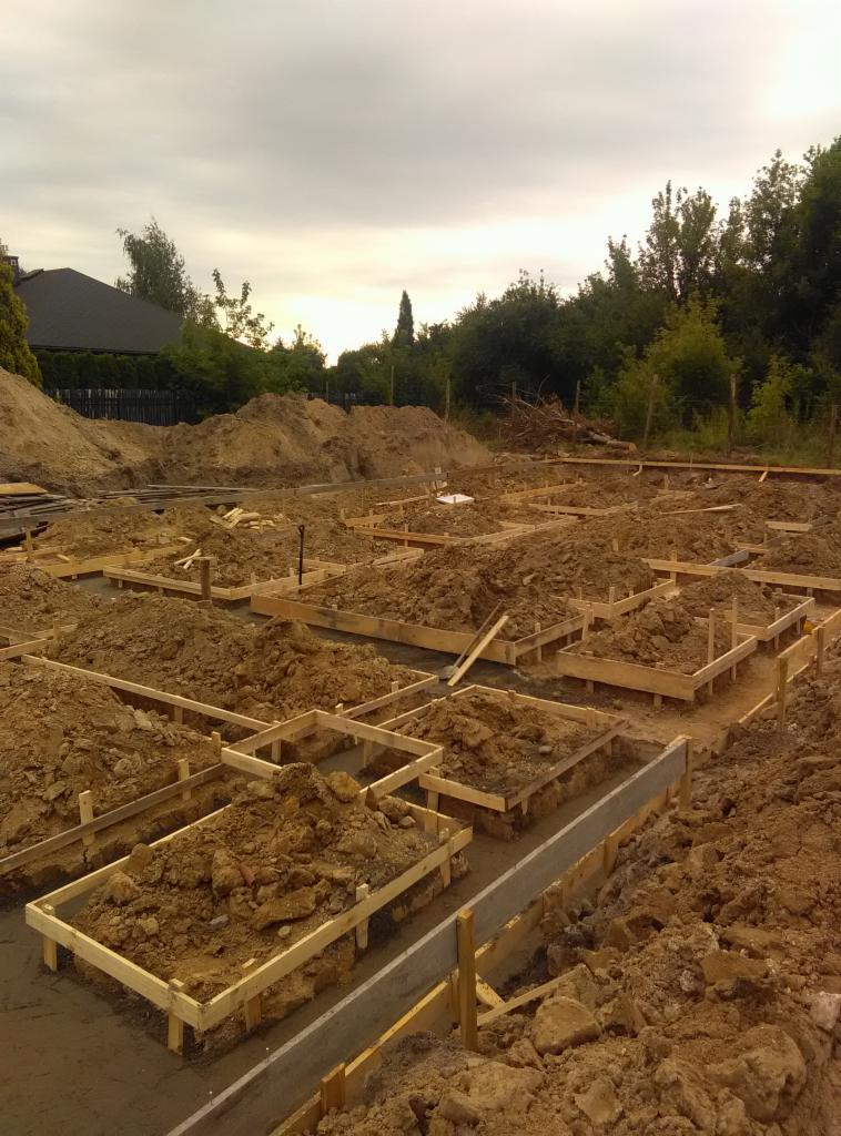Przygotowanie fundamentów pod budowę domu: wykopane i obudowane drewnianymi szalunkami doły wypełnione ziemią, zalane betonem, widoczne zbrojenia i nasypy ziemi w tle.