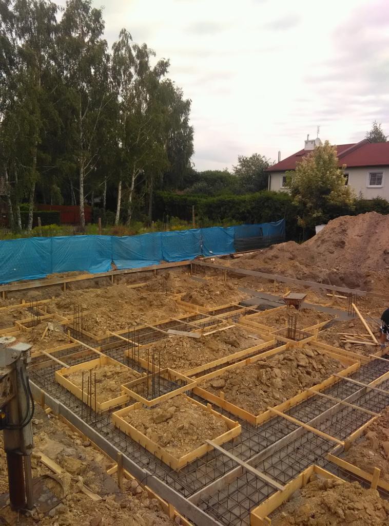 Fundamenty domu murowanego w trakcie budowy: drewniane szalunki wypełnione ziemią i zbrojeniem, widoczna część działki z drzewami i błękitną plandeką w tle.