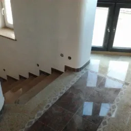 Wykończone schody z brązowymi stopniami i marmurową podłogą z dekoracyjnym wzorem, widoczne gniazdka elektryczne w ścianie, okno z ciemną ramą i fragment kolumny.