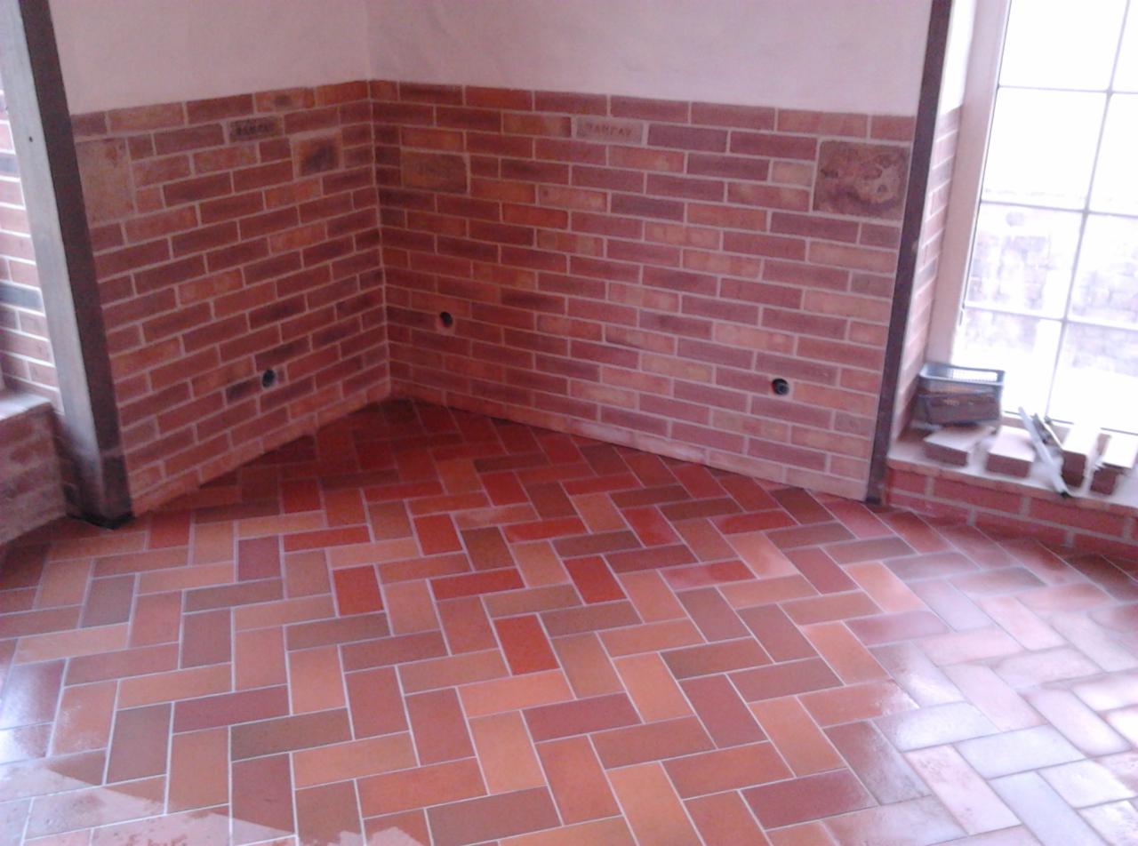 Wykończony fragment pomieszczenia z ceglaną ścianą do połowy wysokości i podłogą z terakoty ułożonej w jodełkę, widoczne otwory na gniazdka elektryczne w ścianie.