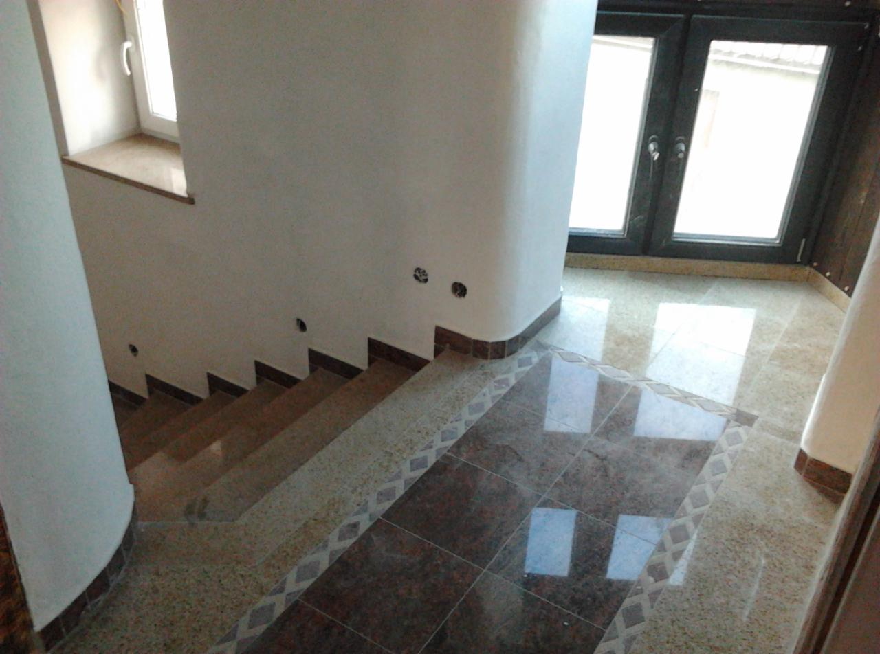 Wykończone schody z brązowymi stopniami i marmurową podłogą z dekoracyjnym wzorem, widoczne gniazdka elektryczne w ścianie, okno z ciemną ramą i fragment kolumny.