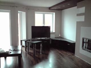 Jasne wnętrze salonu po remoncie z ciemną podłogą, meblami i sufitem podwieszanym. Widoczne duże okna, telewizor na szafce RTV i dekoracyjny kominek.