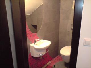 Mała łazienka po remoncie z umywalką na ścianie, muszlą klozetową i szarymi płytkami na ścianach, z dekoracyjnym czerwonym pasem mozaiki od podłogi do lustra.