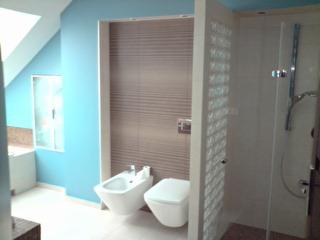 Jasna łazienka na poddaszu z niebieskimi ścianami, białą ceramiką sanitarną (bidet i toaleta) oraz kabiną prysznicową oddzieloną ścianką z luksferów. Na ścianie za ceramiką płytki imitujące lamele.