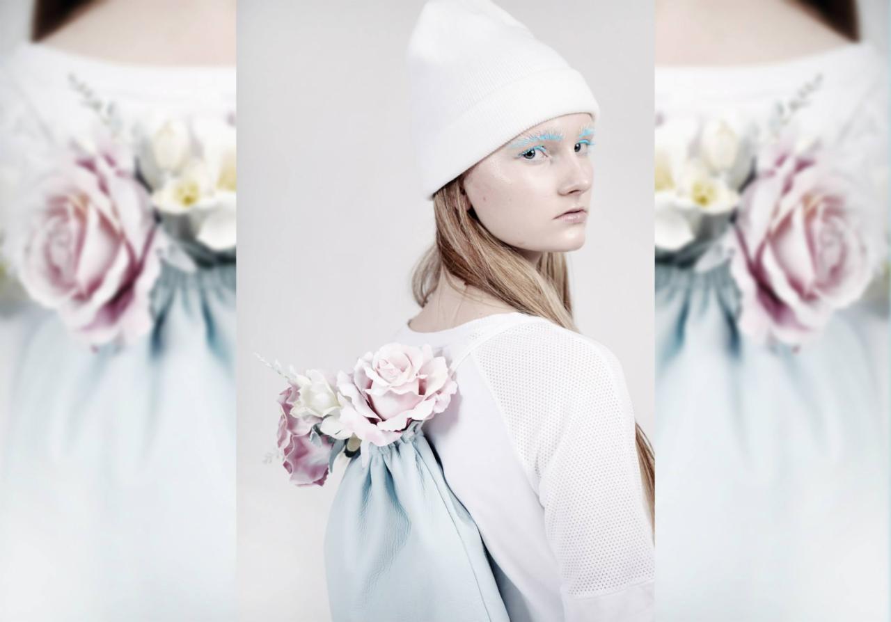 Kobieta z jasnymi brwiami, w białej czapce i koszulce, z jasnoniebieskim plecakiem ozdobionym różowymi i białymi różami, patrzy przez ramię.