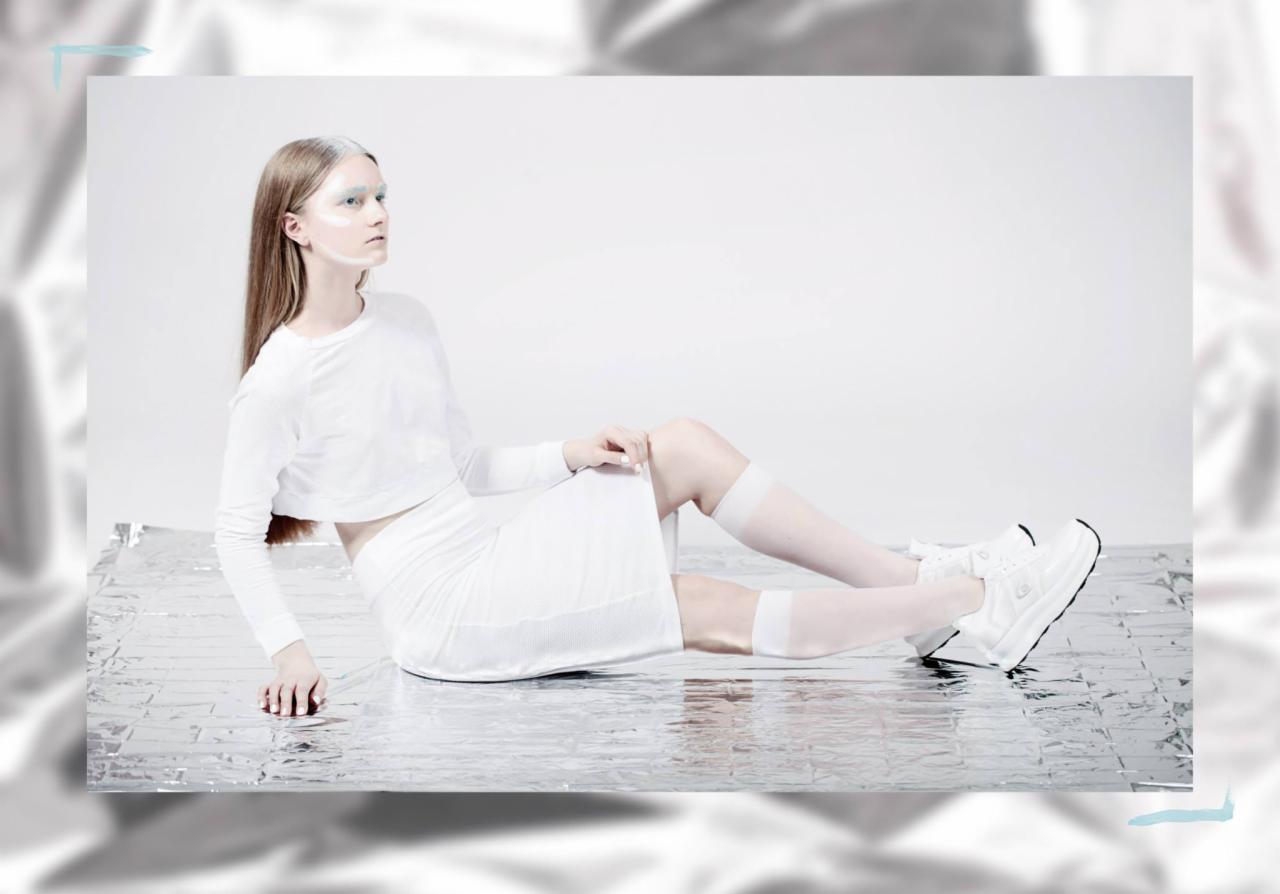 Kobieta w białym stroju pozuje na srebrnej folii, z białym makijażem na twarzy i włosach, patrząc w lewo.