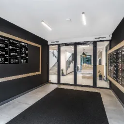 Wnętrze nowoczesnego holu wejściowego do budynku mieszkalnego z czarnymi ścianami, tablicą z logotypami firm, skrzynkami pocztowymi i przeszklonymi drzwiami wejściowymi prowadzącymi na klatkę...