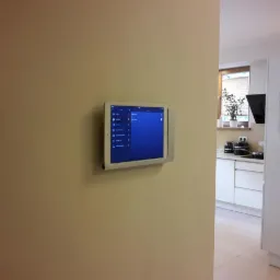 Tablet w srebrnej ramce zamontowany na beżowej ścianie, wyświetlający interfejs systemu zarządzania domem, z widoczną kuchnią w tle.
