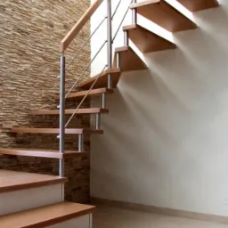 Drewniane schody z metalową balustradą, osadzone przy ścianie wyłożonej dekoracyjnym kamieniem, widoczne z dołu.