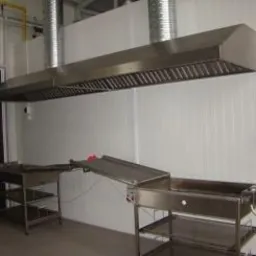 Profesjonalna kuchnia przemysłowa z dużym, metalowym okapem wentylacyjnym, stalowymi stołami roboczymi i gaśnicą na ścianie.