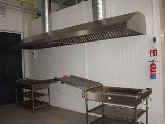 Profesjonalna kuchnia przemysłowa z dużym, metalowym okapem wentylacyjnym, stalowymi stołami roboczymi i gaśnicą na ścianie.