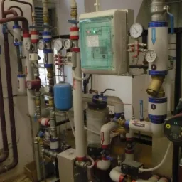 Złożona instalacja grzewcza z licznymi rurami, zaworami, manometrami i pompą obiegową Grundfos, umieszczona w pomieszczeniu technicznym.